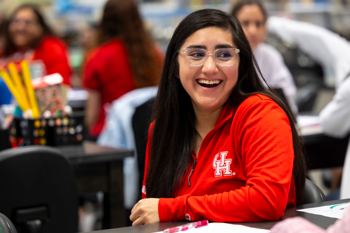 A woman with glasses and a UH t-shirt smiling in a college classroom