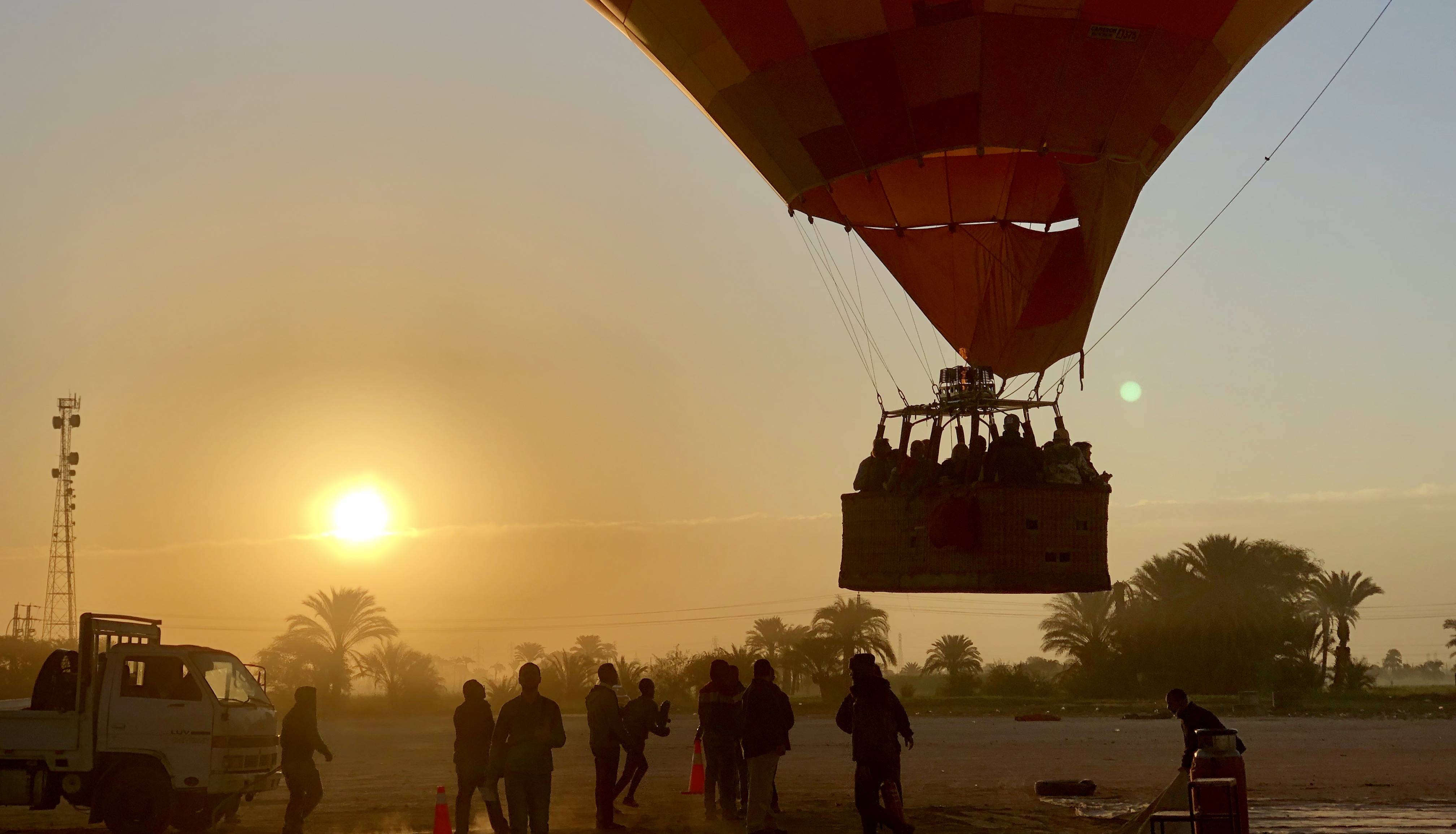 People preparing a hot air balloon for takeoff in front of a sunrise and palm trees