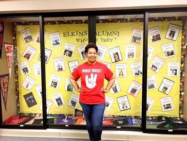 A woman with short brown hair wearing a red shirt standing in front of a high school display case