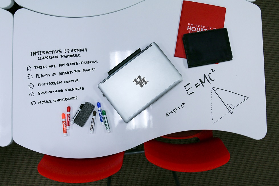 Overhead view of a laptop, notebooks, colored dry-erase markers and an eraser on a white table. with class notes and handouts on top of a white table. The table has additional notes written on it. Notes and diagrams are written on the tabletop in black.