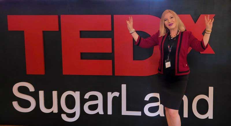 A woman in red business jacket and black dress stands in from of a large sign reading "TEDx Sugar Land"